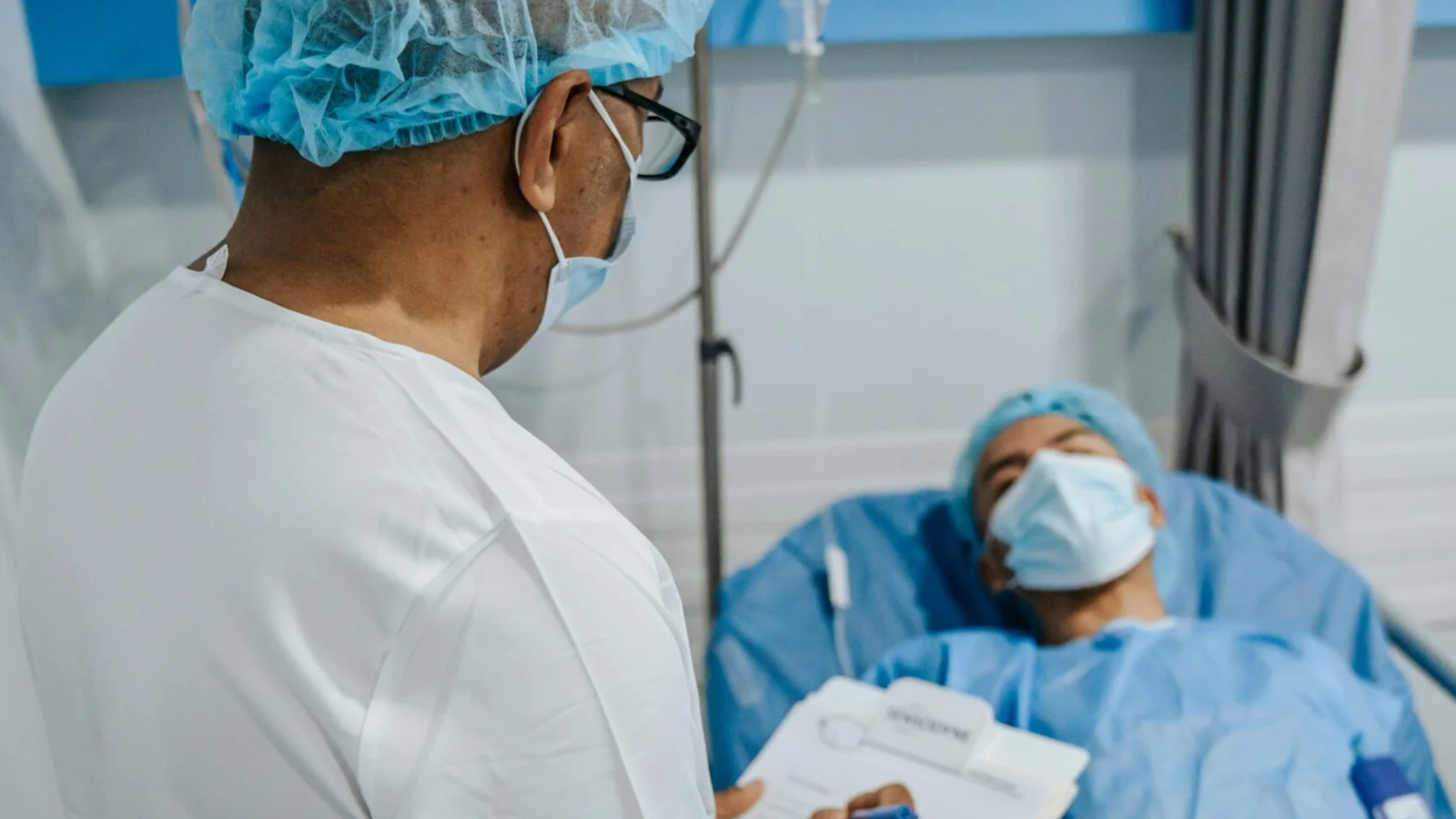 A side view of a medical professional, wearing a white coat, glasses, surgical cap, and mask, speaking to a patient lying in a hospital bed. The patient, also wearing a surgical cap and mask, is blurred in the background, suggesting the focus is on the doctor's action. The doctor is holding a clipboard.