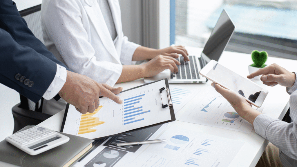 A close-up of a business meeting where a man in a suit points to a bar chart on a clipboard, while two colleagues look on, using a laptop and a tablet filled with data graphs.