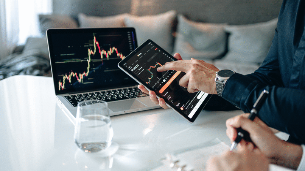 A businessman points to a financial candlestick chart on a tablet, sitting at a desk with a laptop also displaying stock market data, while a colleague takes notes.