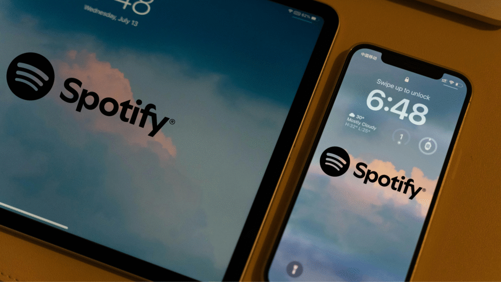 A close-up view of a tablet and a smartphone displaying the Spotify logo over a cloudy sky background, emphasizing music consumption across multiple mobile devices.