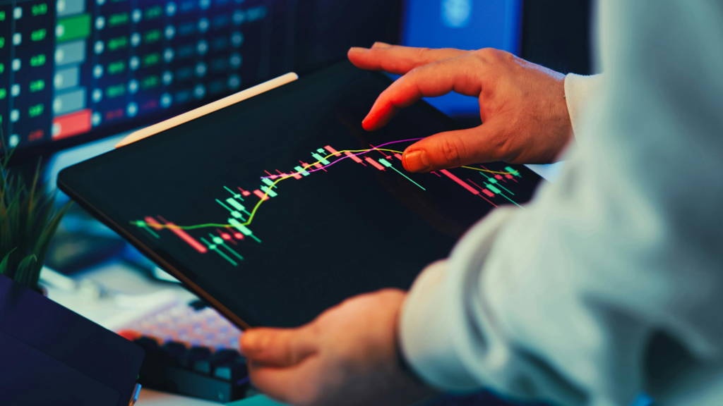 A person holding a tablet displaying a colorful candlestick trading chart with trend lines, with a computer monitor showing additional financial data in the background.